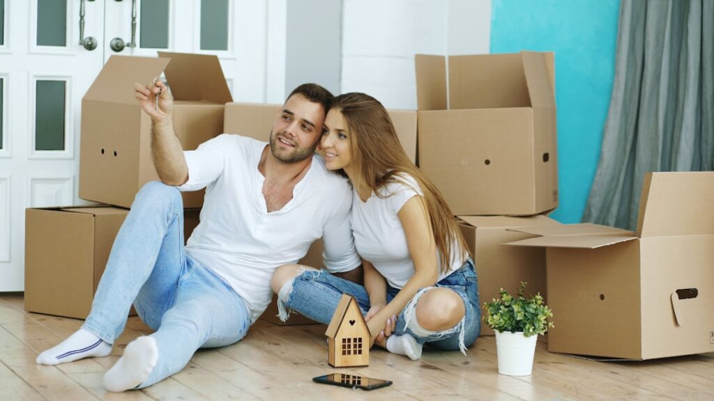 Happy couple sitting on floor with moving boxes and a miniature house model holding keys representing a life-stage housing transition